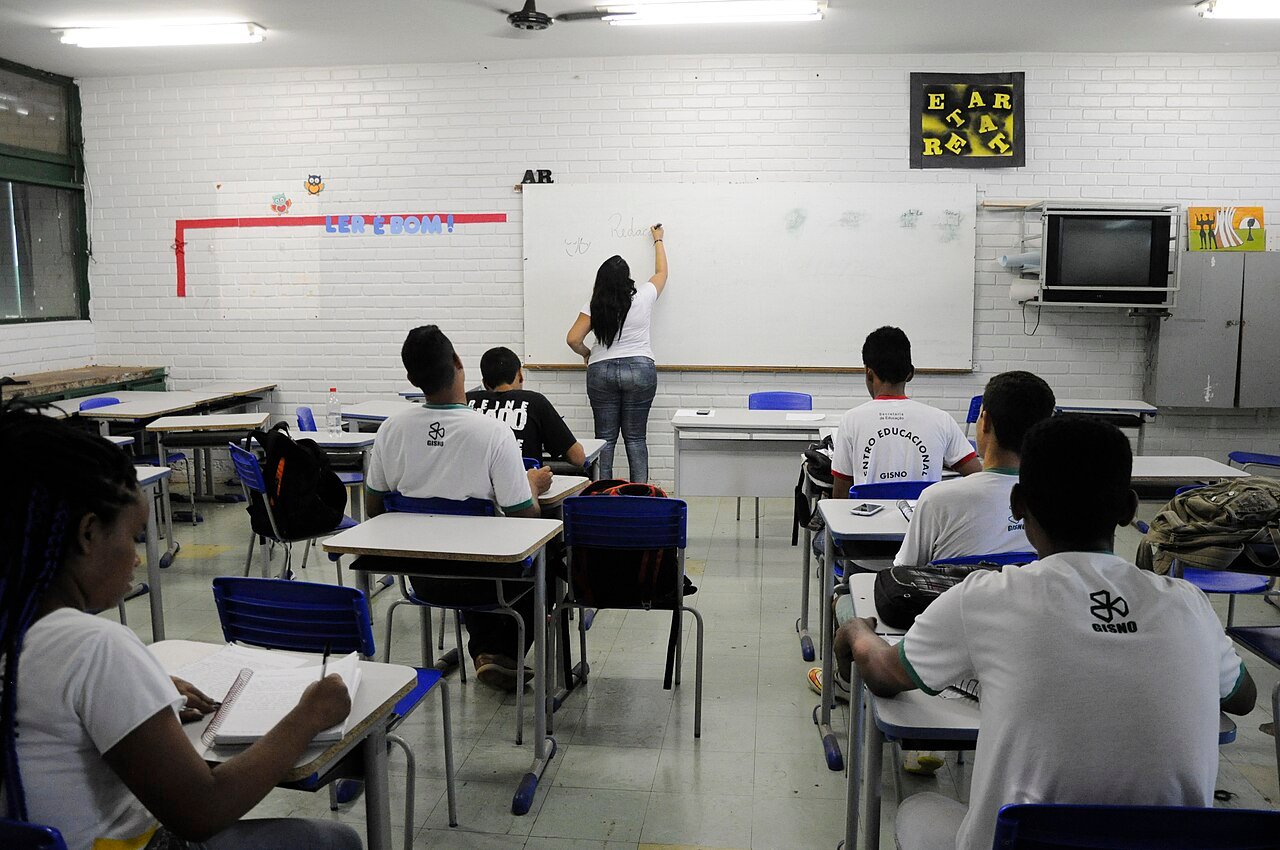 Sala de aula de escola pública do Distrito Federal, com alunos de costas e professora escrevendo no quadro