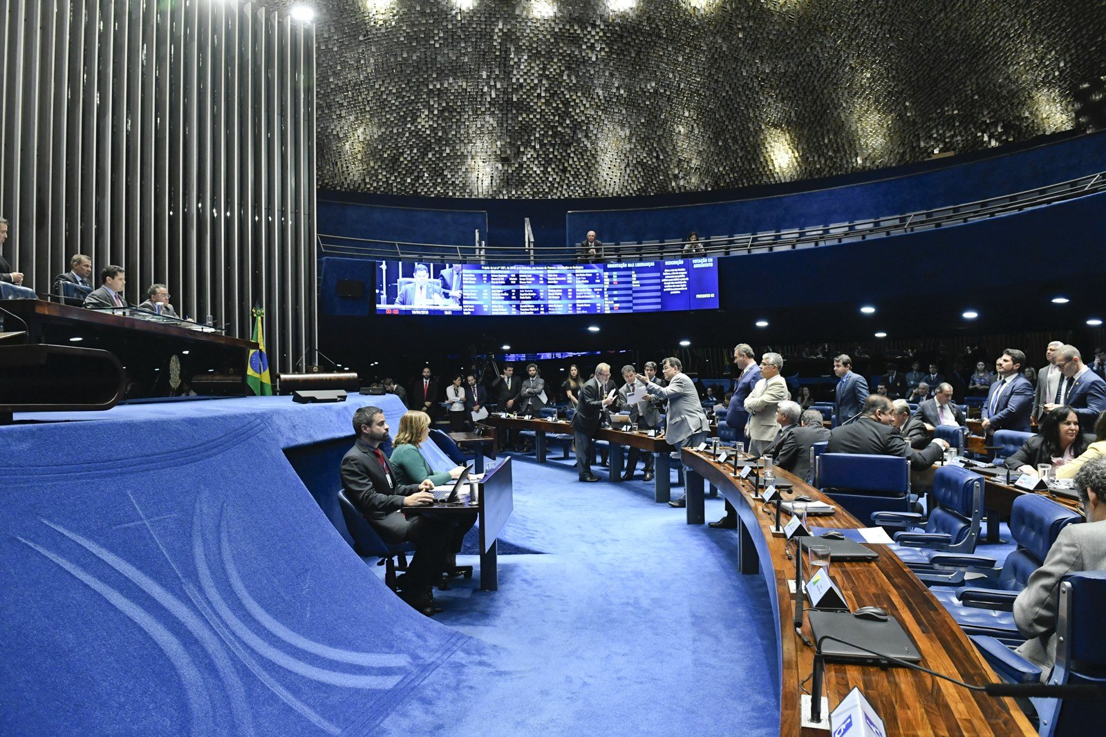 Plenário do Senado Federal em sessão deliberativa, com painel de votação ao centro e senadores em pé entre as bancadas