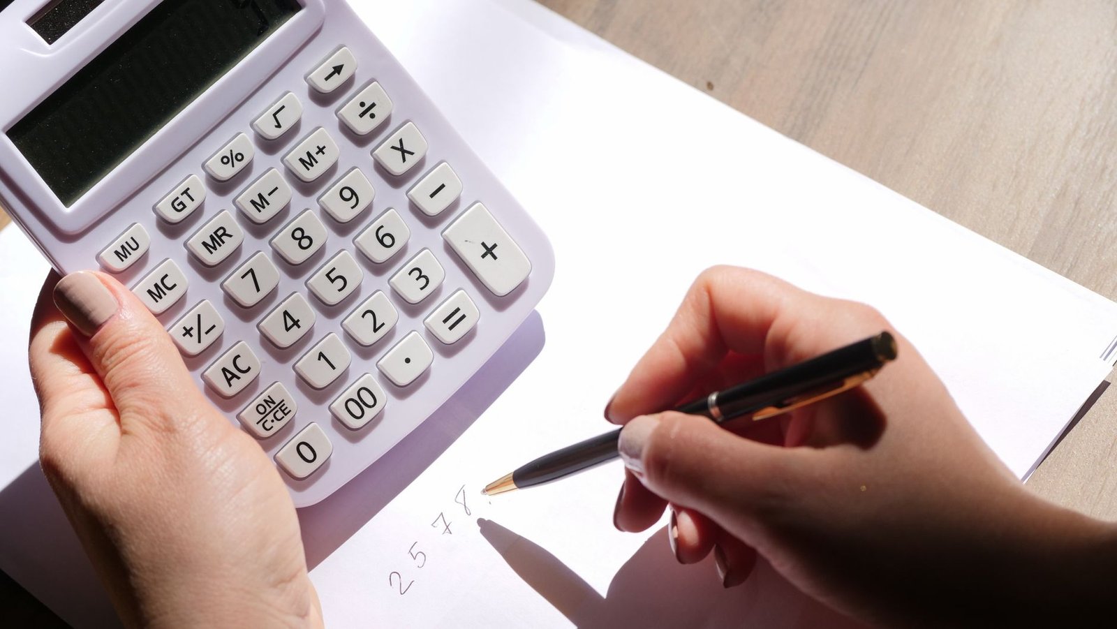 Mãos de uma pessoa usando calculadora e escrevendo números em uma folha sobre a mesa