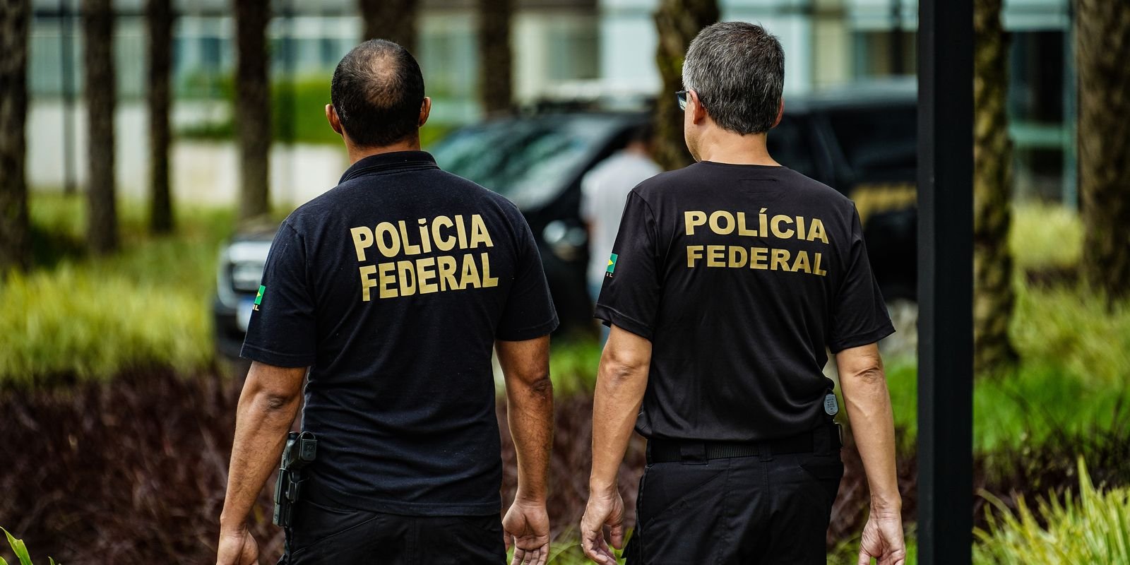 Dois agentes da Polícia Federal caminham de costas, com a inscrição Polícia Federal nas camisas