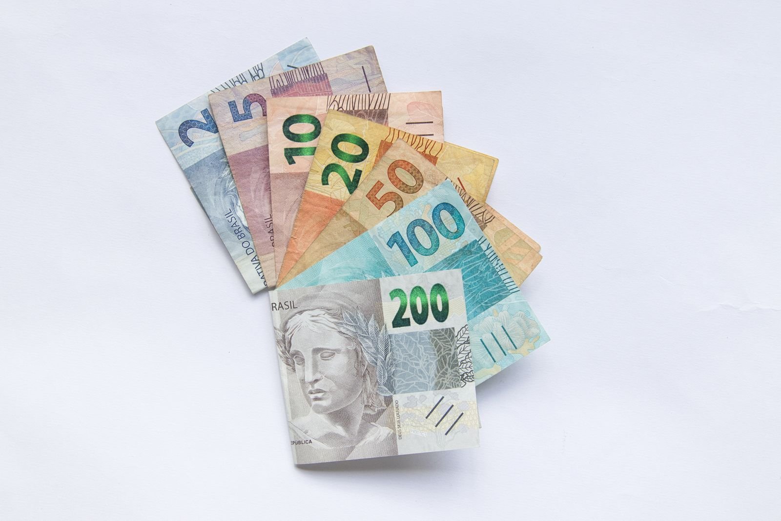 Cédulas de real de 2, 5, 10, 20, 50, 100 e 200 dispostas em leque sobre fundo branco