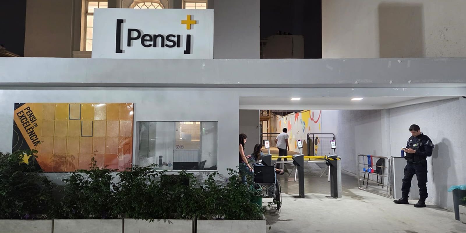 Entrada do Colégio Pensi, no Flamengo, no Rio de Janeiro, à noite, com catracas e um policial na porta