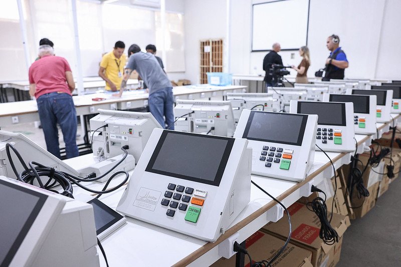 Urnas eletrônicas enfileiradas em sala de preparação da Justiça Eleitoral com técnicos ao fundo