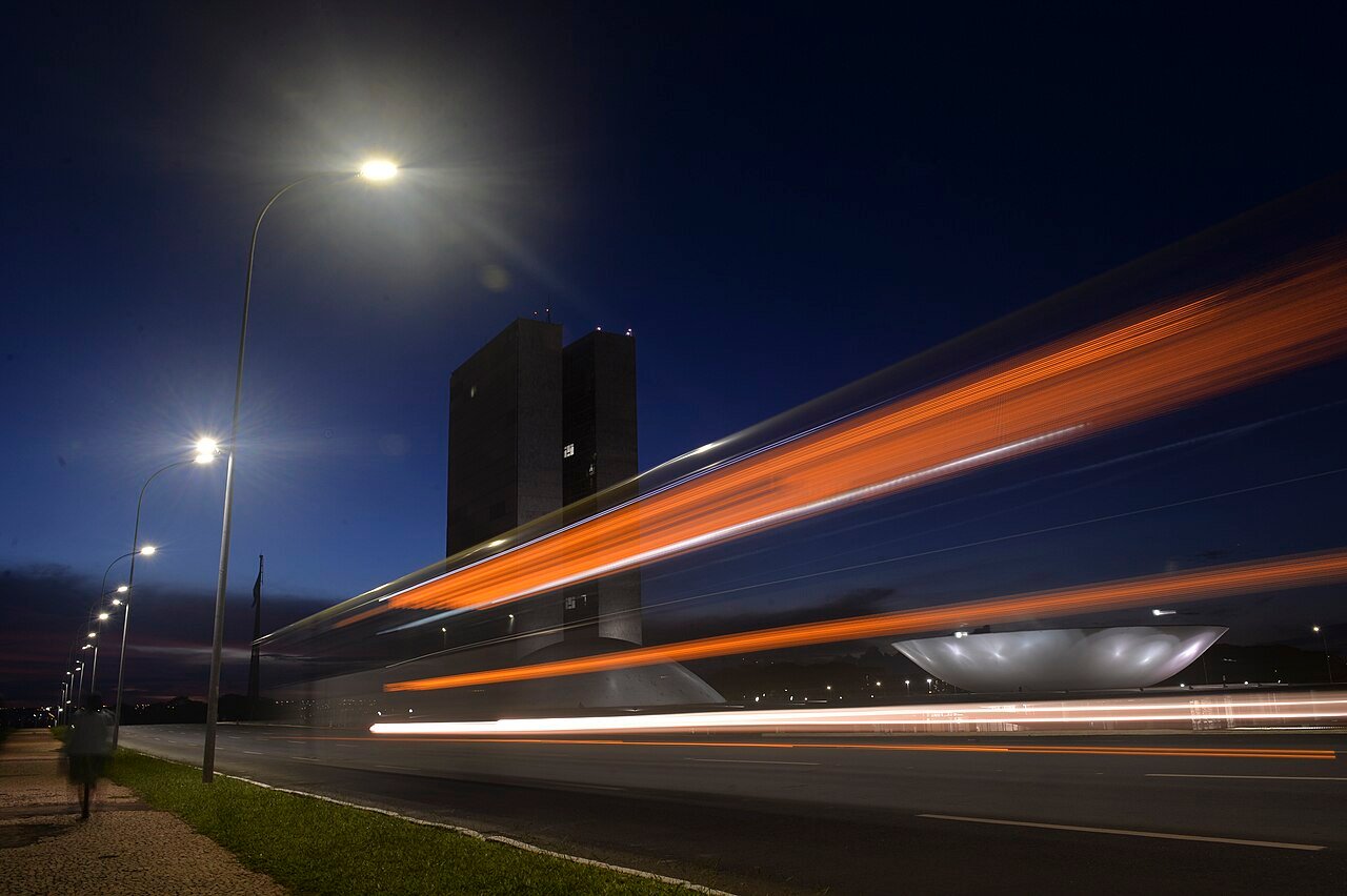 Torres e cúpulas do Congresso Nacional, em Brasília, ao entardecer, com rastros de luz de veículos na pista em primeiro plano