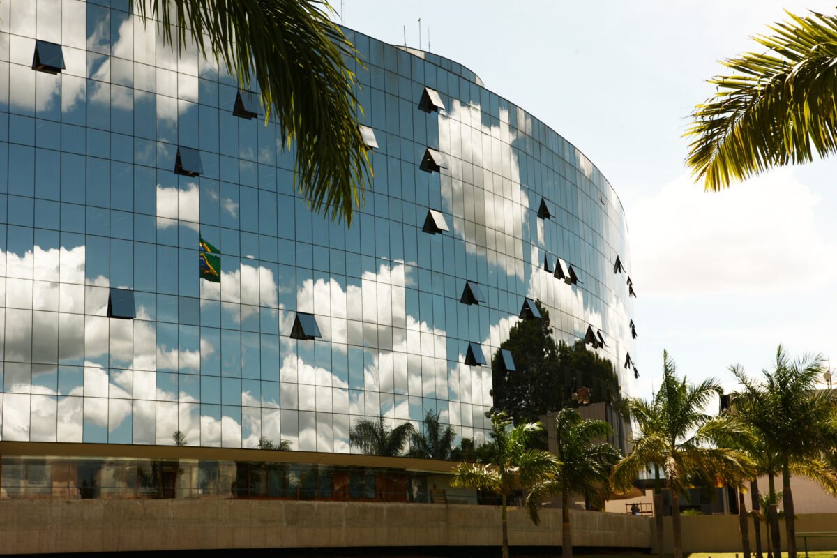 Fachada de vidro do Anexo II do Supremo Tribunal Federal, em Brasília, com a bandeira do Brasil refletida e palmeiras em primeiro plano