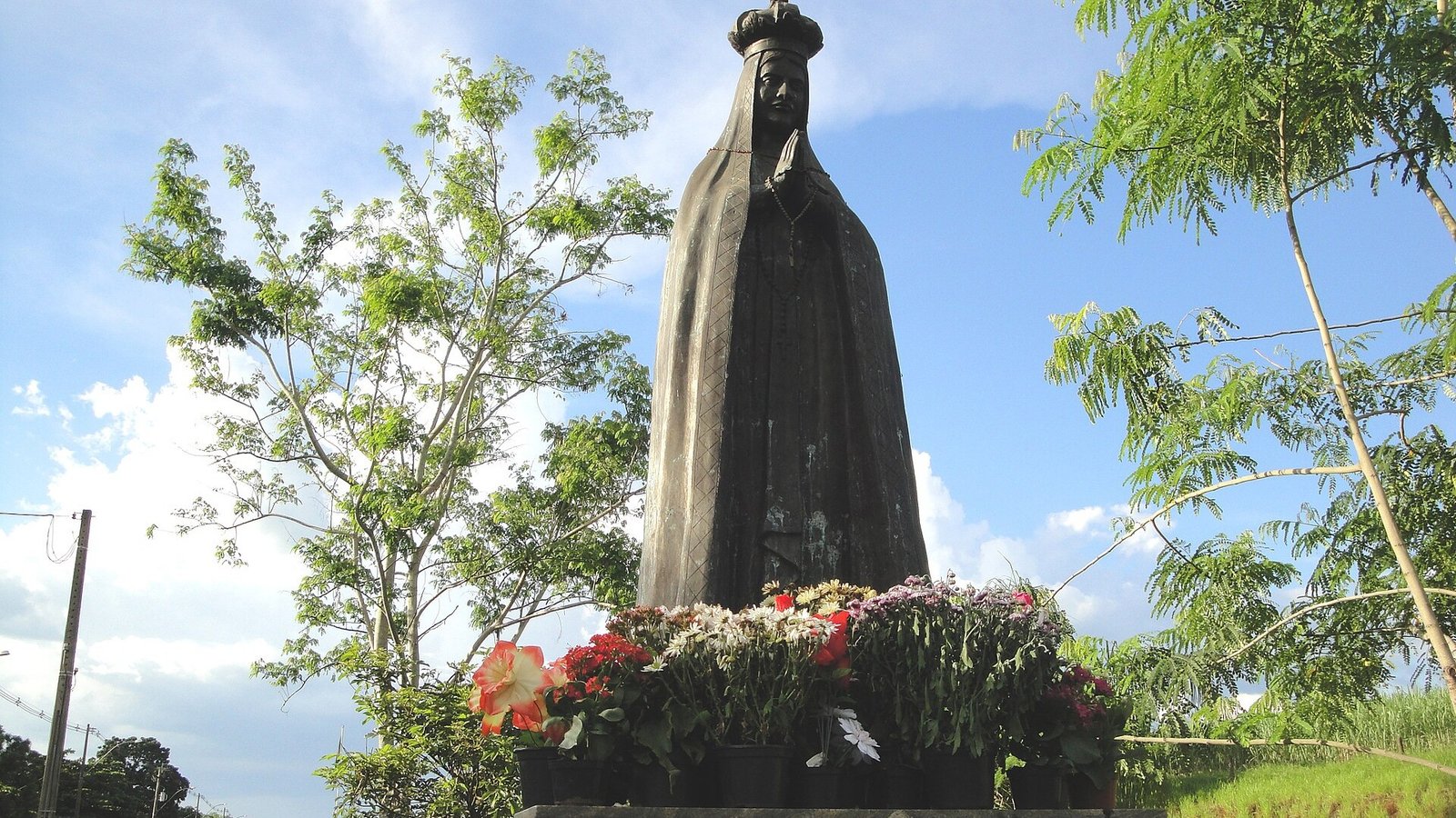 Estátua de Nossa Senhora Aparecida cercada de vasos de flores, à beira de uma rodovia, sob céu azul