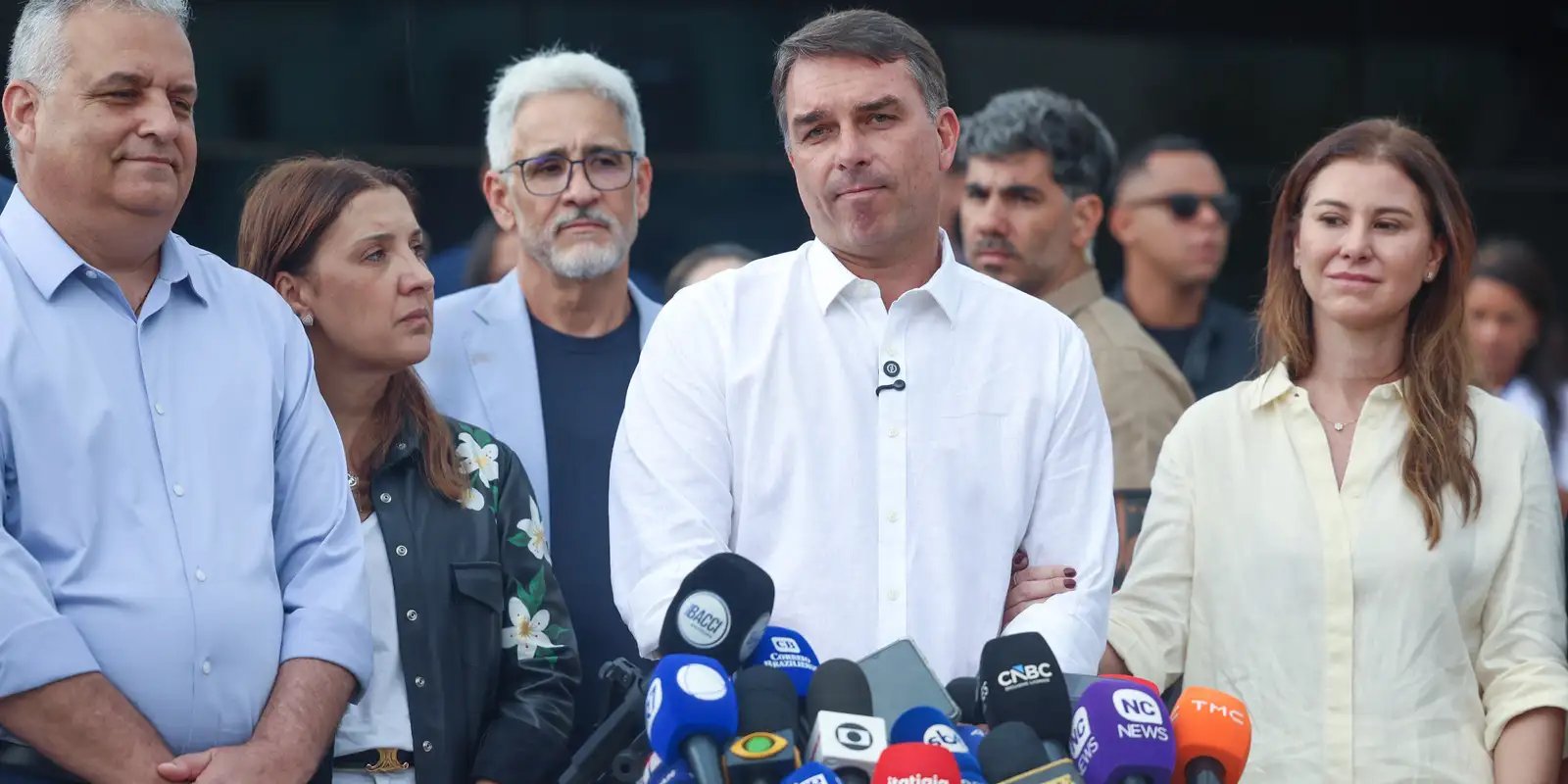 O senador Flávio Bolsonaro, de camisa branca, diante de microfones durante entrevista coletiva, cercado de aliados