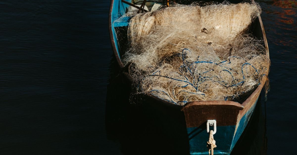 Barco de pesca de madeira azul com rede de pesca amontoada, em águas escuras (imagem ilustrativa)