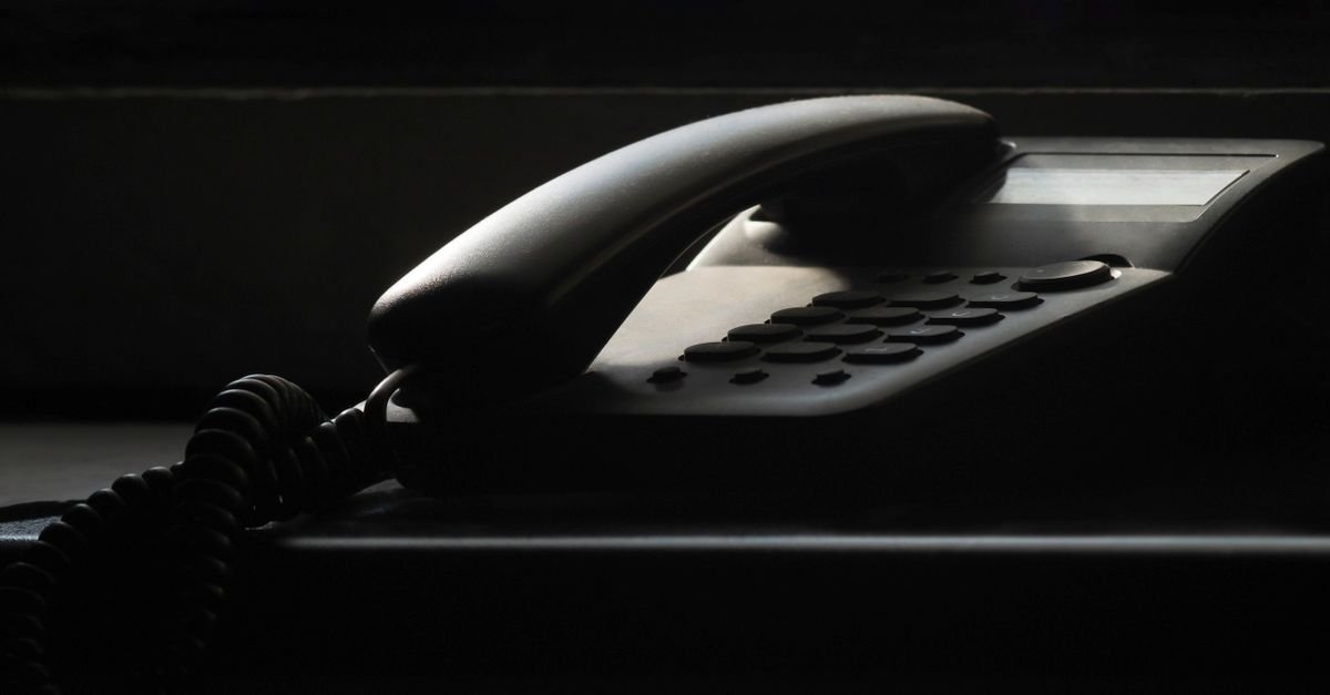 Telefone fixo de mesa fotografado em contraluz, com o fone no gancho e o fio espiralado