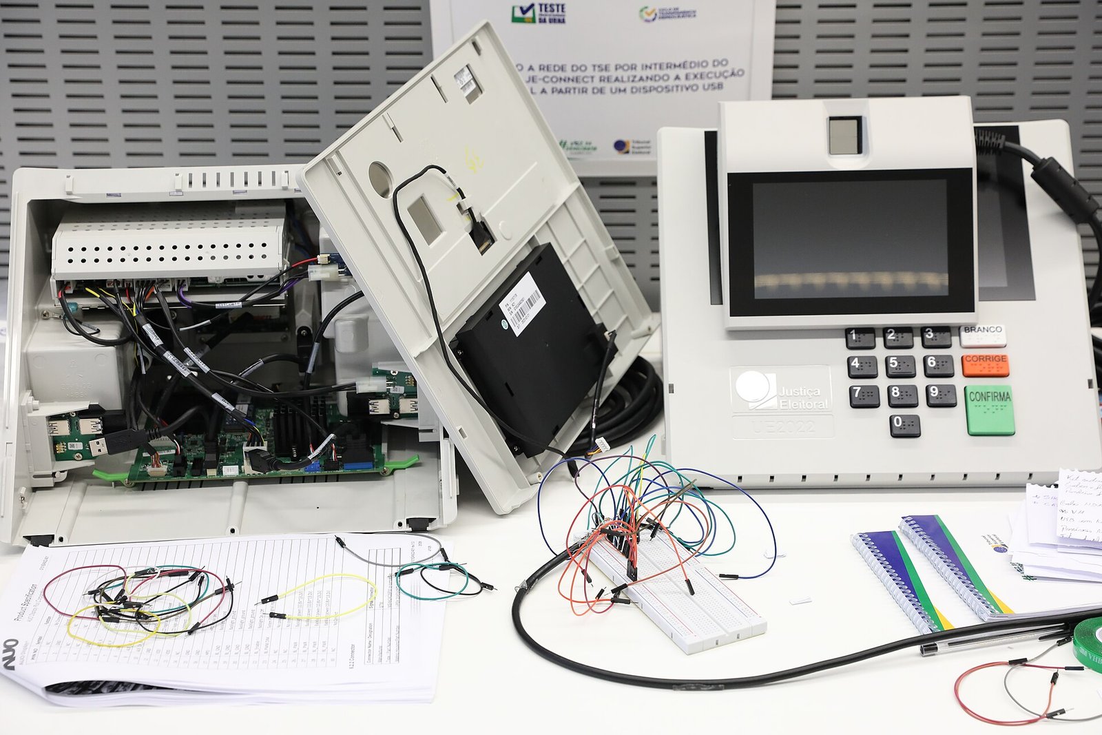Urna eletrônica da Justiça Eleitoral aberta sobre bancada, com placa de circuito e conexões expostas, ao lado de outra urna fechada com o teclado numérico