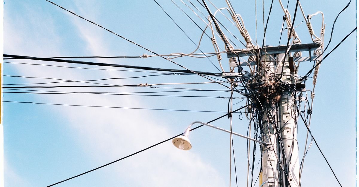 Poste urbano com emaranhado de fios e luminaria, visto de baixo contra o ceu azul