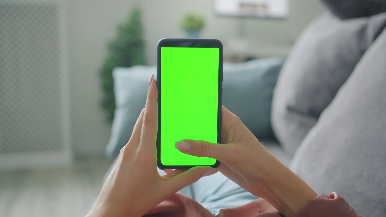 Mãos seguram um celular com a tela verde, em sala com sofá
