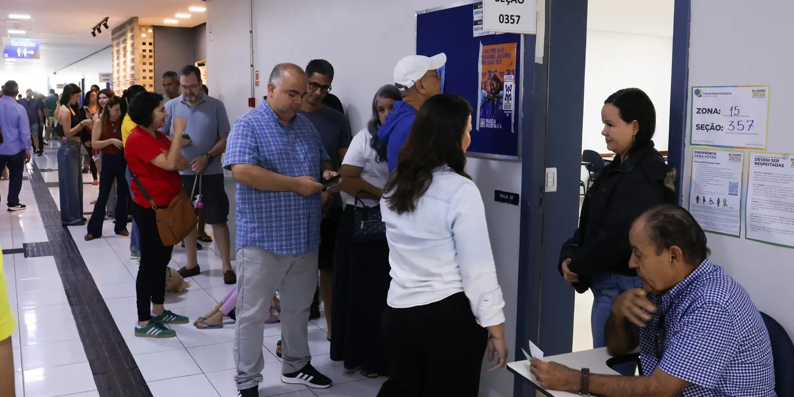 Eleitores em fila diante de uma seção eleitoral identificada por zona e número no primeiro turno de 2026