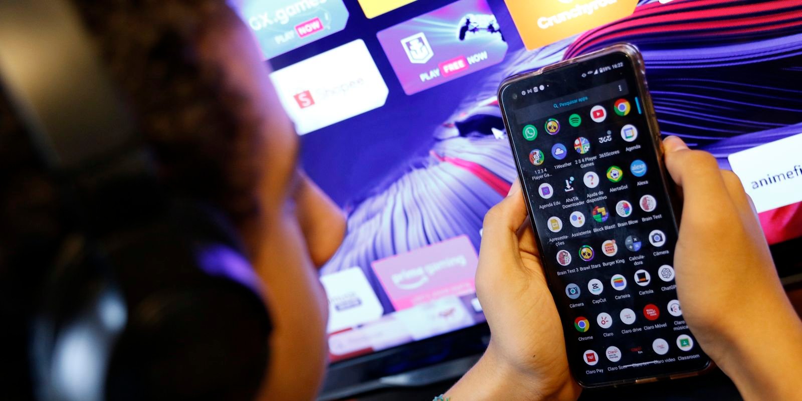 Criança de costas segura celular com tela de aplicativos diante de uma televisão ligada