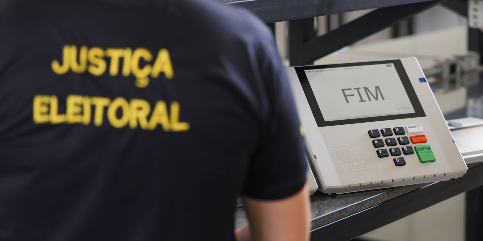Servidor da Justiça Eleitoral de costas ao lado de uma urna eletrônica com a tela mostrando a palavra FIM
