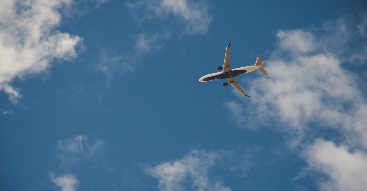 Avião comercial em voo visto de baixo, com céu azul e nuvens ao fundo
