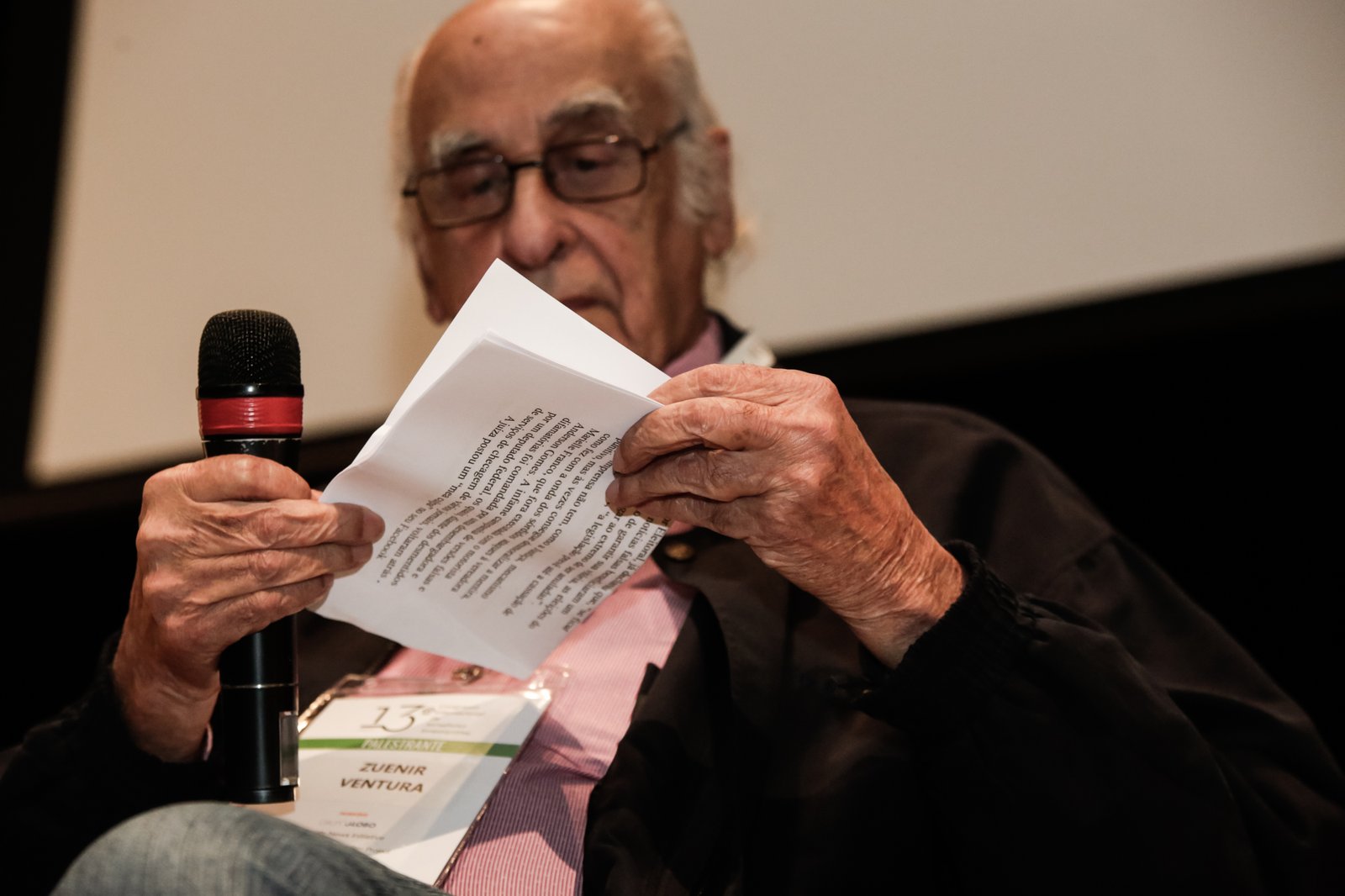 Zuenir Ventura lê uma folha impressa segurando o microfone, com o crachá de palestrante do 13º Congresso Internacional de Jornalismo Investigativo no colo